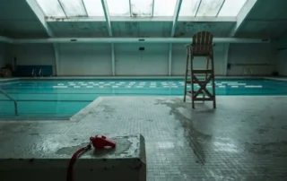 Verlassenes Hallenbad im Winter mit türkisfarbenem Wasser, Bahnseilen, leerem Rettungsstuhl und einer Pfeife auf den nassen Fliesen.