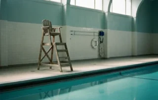 Leerer Rettungsstuhl neben einem ruhigen Hallenbad mit türkisblauem Wasser und Rettungsausrüstung an der gekachelten Wand.