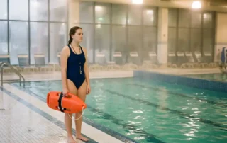 Junger Rettungsschwimmer-Azubi in Badebekleidung hält Rettungsring am Beckenrand einer Hallenbad-Anlage.