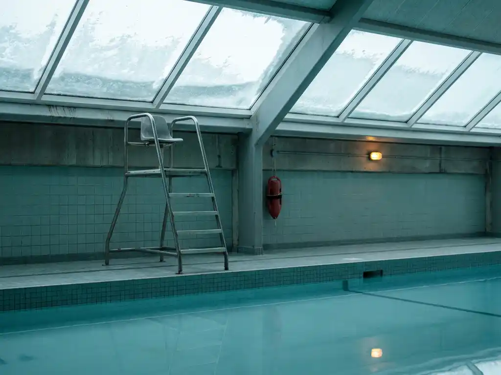 Leerer Rettungsstuhl an einem verlassenen Hallenbad im Winter, frostige Oberlichter, stilles blaues Wasser, roter Rettungsring an der Wand.