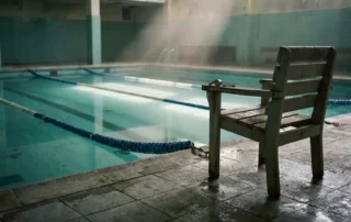 Verlassenes Hallenbad mit türkisfarbenem Wasser, Bahnseilen und einem leeren Rettungsstuhl, auf dem eine Pfeife liegt.