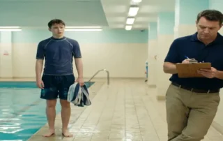 Zeitarbeiter in marineblauer Badebekleidung steht unsicher am Rand eines Hallenbades, während ein Manager ein Klemmbrett prüft.