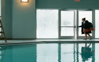 Leerer Rettungsstuhl an einem Hallenbad im Winter, während ein neuer Bademeister durch Milchglastüren mit Ausrüstung eintritt.