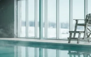 Leerer Rettungsstuhl an einem menschenleeren Hallenbad mit dampfendem türkisfarbenem Wasser und Winterfrost hinter Panoramafenstern.