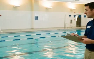 Zertifizierter Poolprofi mit Klemmbrett am Beckenrand, Rettungsschwimmer auf erhöhtem Stuhl im Hintergrund, Hallenbad mit türkisblauem Wasser.
