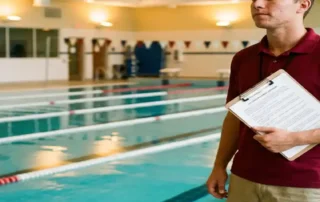 Rettungsschwimmer in roter Uniform prüft Dokument auf Klemmbrett am Beckenrand eines Indoor-Schwimmbads.
