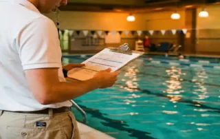 Rettungsschwimmer in Uniform prüft Dienstplan auf Klemmbrett am Rand eines Hallenbades mit türkisfarbenem Wasser.