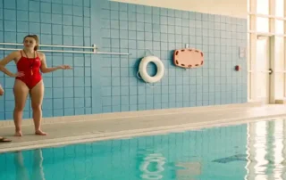 Rettungsschwimmerin in rotem Badeanzug schult neues Zeitpersonal am Beckenrand einer Hallenbad-Anlage mit Rettungsausrüstung.