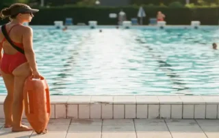 Rettungsschwimmer im roten Badeanzug am Beckenrand eines öffentlichen Freibades mit Rettungsring, der das Wasser beobachtet.