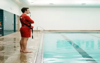 Rettungsschwimmer in roter Badebekleidung überwacht ruhiges Hallenbad, Rettungsausrüstung an türkisfarbener Kachelwand befestigt.