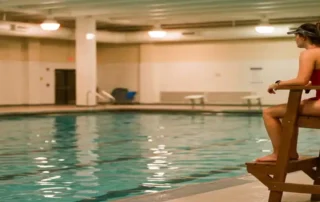 Rettungsschwimmerin in rotem Badeanzug auf erhöhtem Beobachtungsstuhl überwacht ein ruhiges Hallenbad mit türkisfarbenem Wasser.