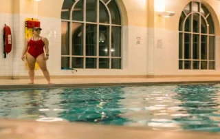 Rettungsschwimmerin in roter Uniform überwacht aufmerksam das türkisfarbene Wasser einer Hallenbad-Anlage, Rettungsausrüstung im Hintergrund.