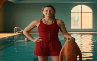 Rettungsschwimmer in roter Uniform steht am Beckenrand eines Hallenbades, Hände auf Rettungsboje, türkisblaues Wasser im Hintergrund.