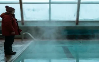 Bademeister in roter Winterjacke bewacht leeres Hallenbad mit dampfendem Wasser und frostbedeckten Oberlichtern im Winter.
