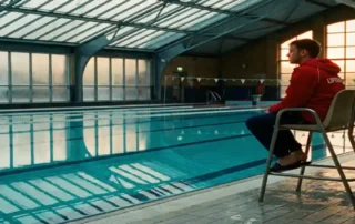 Rettungsschwimmerin in rotem Badeanzug überwacht leeres Hallenbad mit türkisblauem Wasser und Glasdecke im Winter.