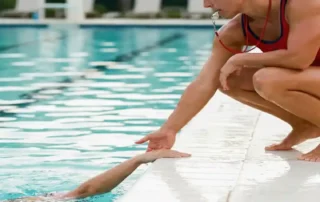 Rettungsschwimmer kauert am Beckenrand und hilft einem Schwimmer, türkisfarbenes Wasser glänzt im Hintergrund.
