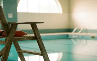Leerer Rettungsschwimmer-Stuhl mit aufgerolltem Rettungsring an einem ruhigen Hallenbad mit türkisfarbenem Wasser.