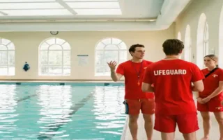 Rettungsschwimmer in roter Uniform gibt Teambriefing am Beckenrand einer hellen Hallenbad-Anlage mit türkisblauem Wasser.