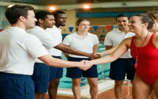 Rettungsschwimmerin in roter Uniform wird von ihrem neuen Team am Beckenrand einer Hallenbadanlage herzlich begrüßt.