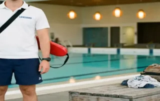 Rettungsschwimmer in Uniform mit rotem Rettungsring am Beckenrand eines modernen Hallenbades, türkisfarbenes Wasser im Hintergrund.