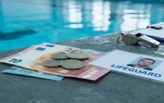 Euro-Scheine und Münzen neben einem Rettungsschwimmer-Ausweis auf nassem Beckenboden, Pfeife und Dienstplan im Hintergrund.