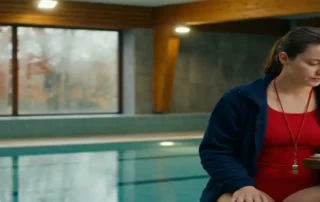 Rettungsschwimmerin in roter Uniform prüft Saisonplan auf Klemmbrett am Hallenbad, Herbstblätter im beschlagenen Fenster sichtbar.