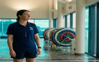 Aquatik-Fachkraft in marineblauer Uniform steht selbstbewusst am Beckenrand einer modernen Hallenschwimmhalle mit türkisfarbenem Wasser.